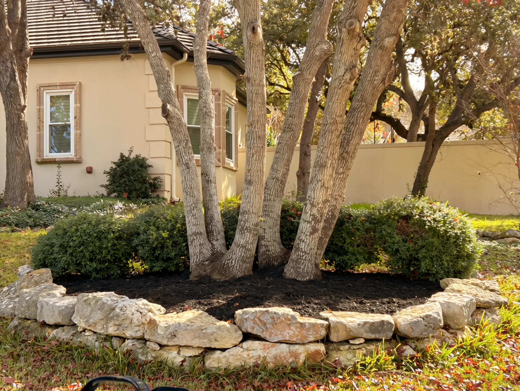 Mortared stone retaining wall around oak with stucco home behind